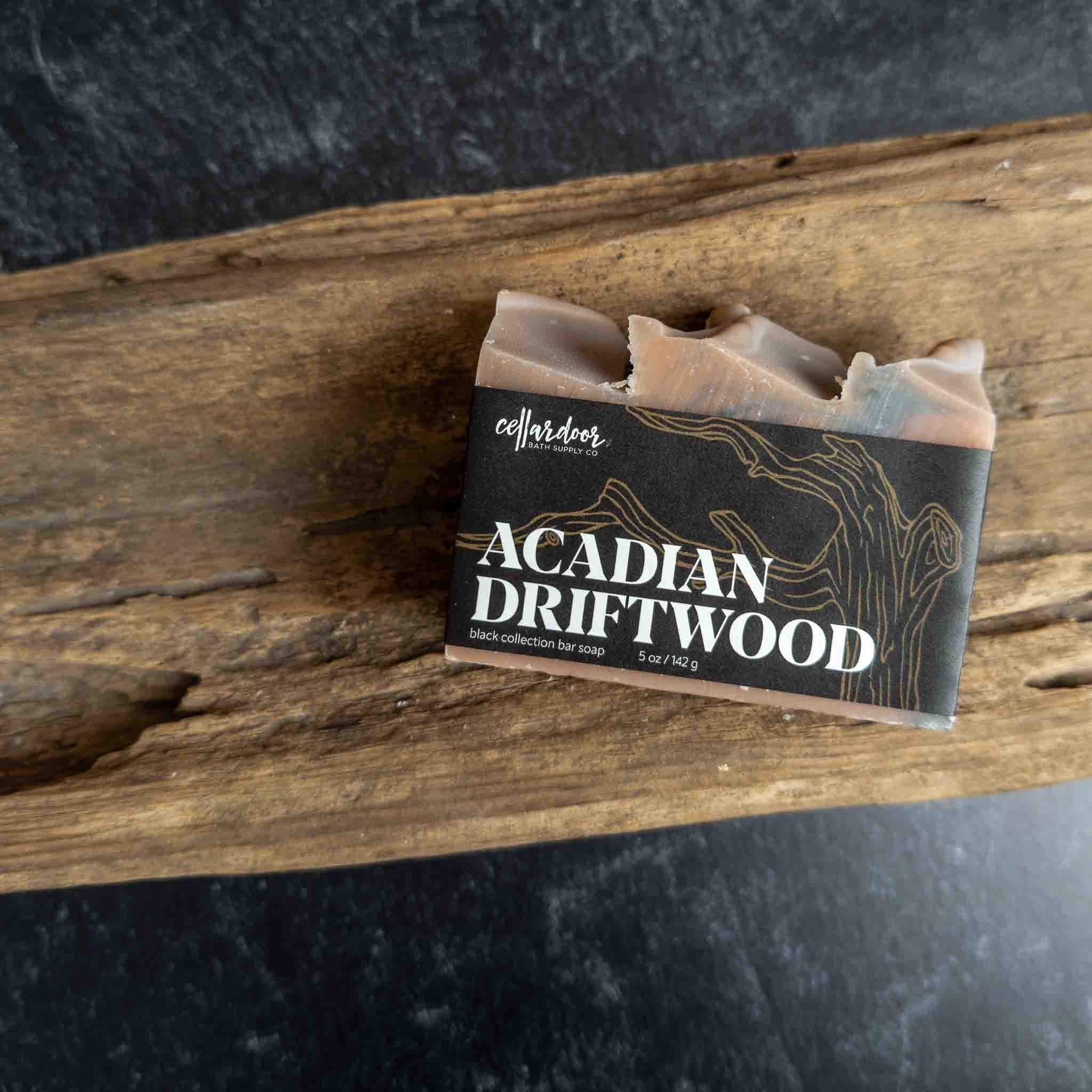 A bar of Collardier Acadian Driftwood soap resting on a rustic piece of wood, with a black label featuring white text and gold driftwood line art.