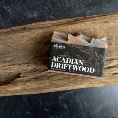 A bar of Collardier Acadian Driftwood soap resting on a rustic piece of wood, with a black label featuring white text and gold driftwood line art.