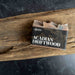 A bar of Collardier Acadian Driftwood soap resting on a rustic piece of wood, with a black label featuring white text and gold driftwood line art.
