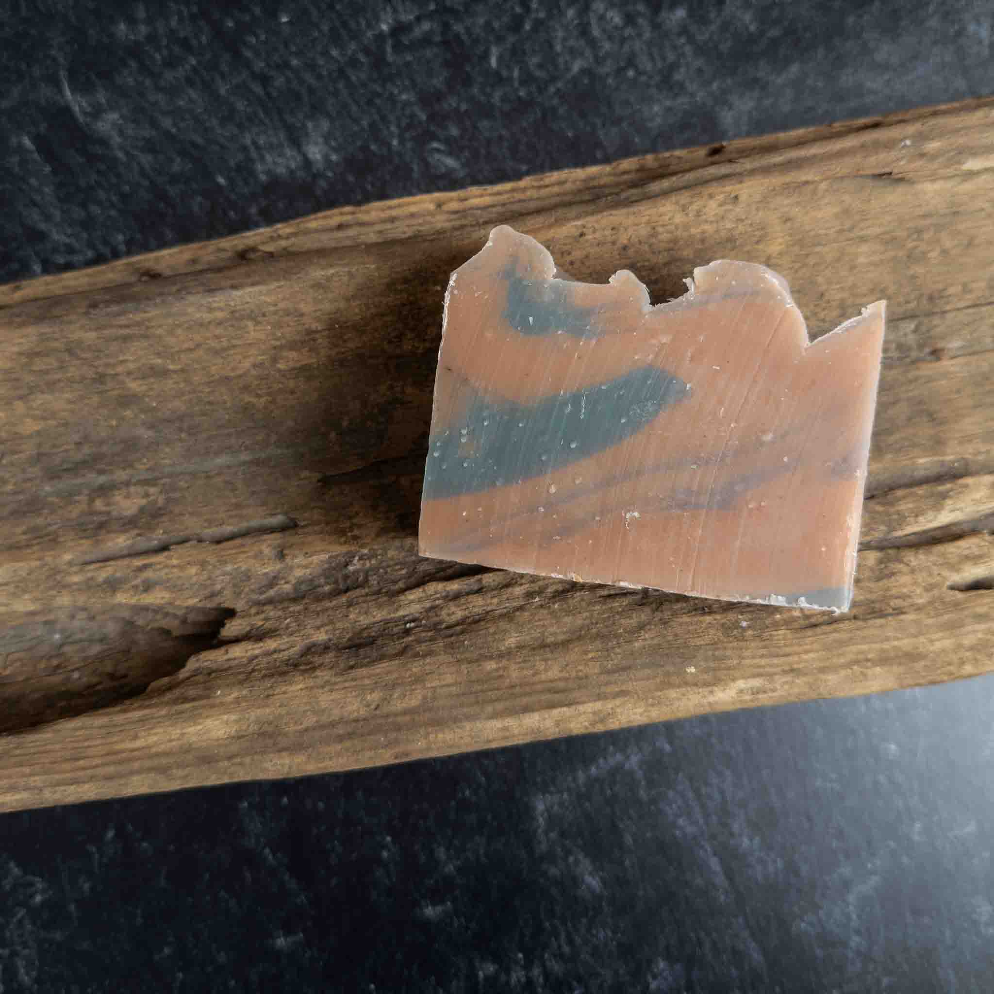 A rectangular bar of handmade soap with a marbled pattern of peach, orange, and slate blue, resting on a piece of weathered driftwood over a dark textured surface.