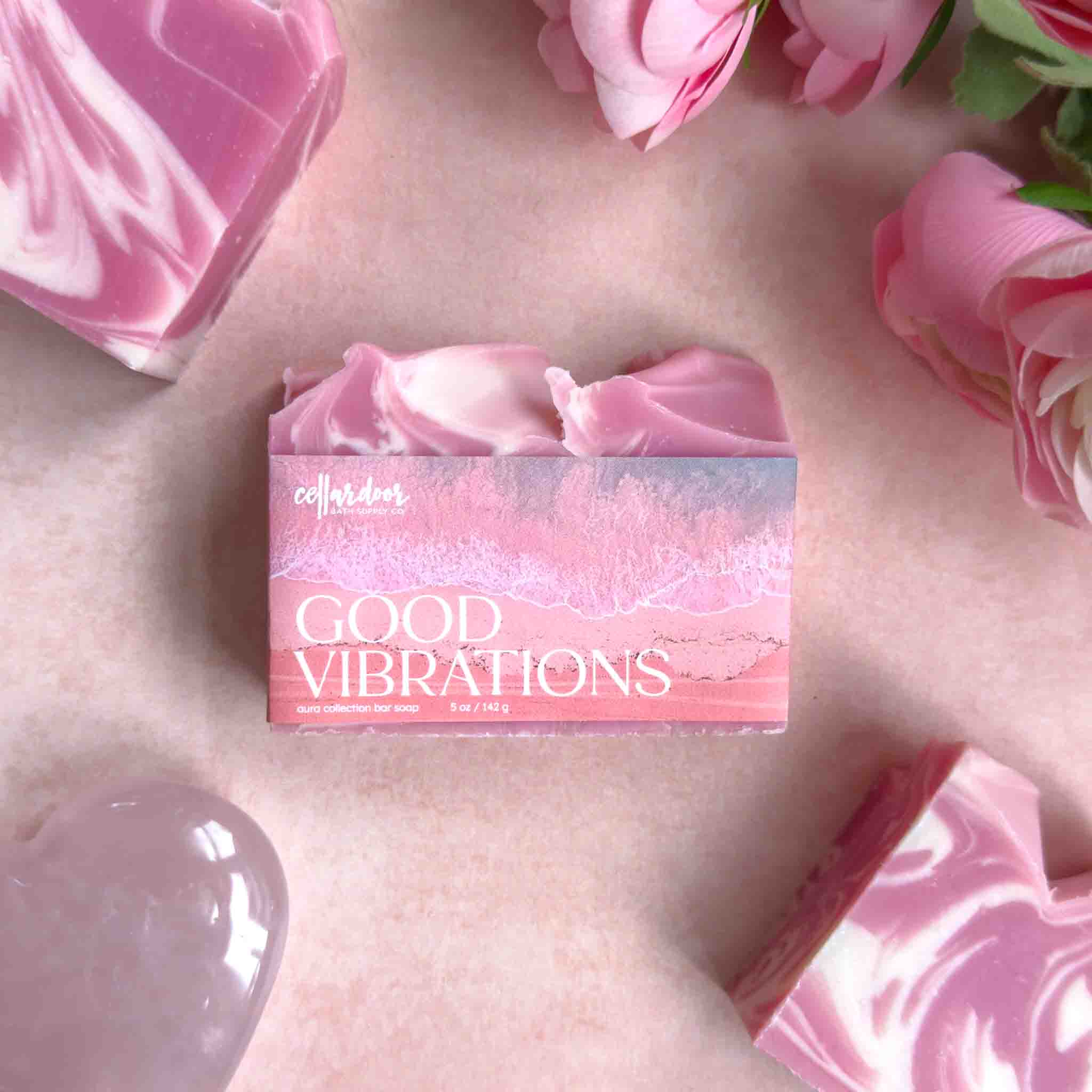 Bar of soap with 'Good Vibrations' packaging on a surface with pink flowers and soap.