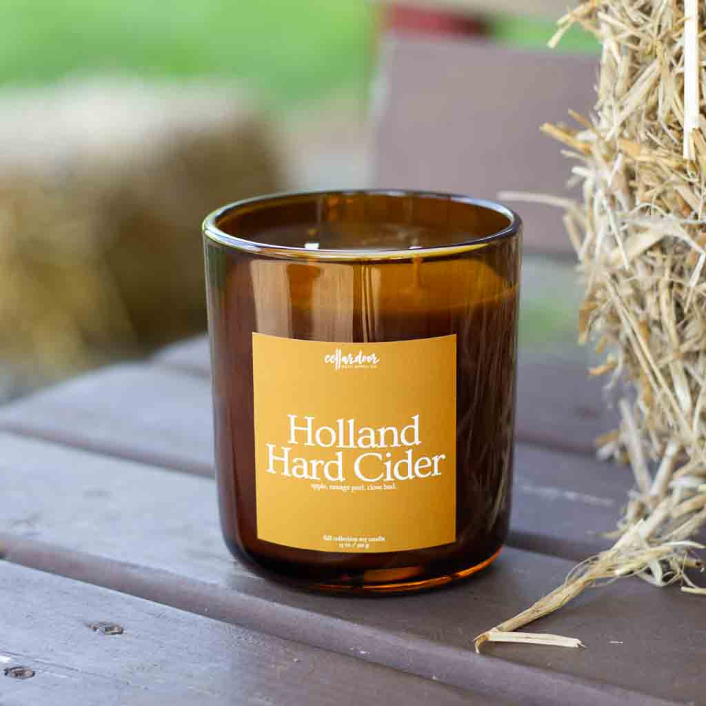 An amber jar candle labeled 'Holland Hard Cider' sits on a brown wood bench beside a straw bale.