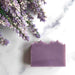 A rectangular bar of lavender-colored soap with rustic, unevenly broken edges, styled on a white marble surface next to dried lavender flowers.