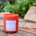 An orange soy candle with a label reading 'Lavender Pumpkin', placed on a brick surface with green plants in the background.