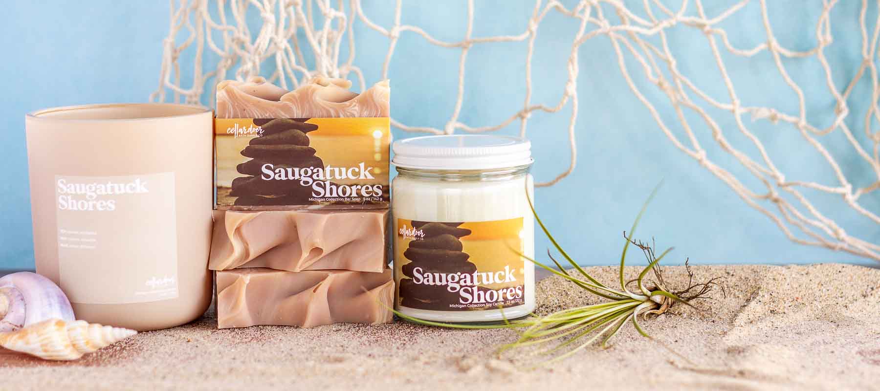 A collection of soaps and candles named Saugatuck Shores rest on a bed of sand.