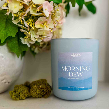 Candle labeled 'Morning Dew' with flowers and decorative elements in the background