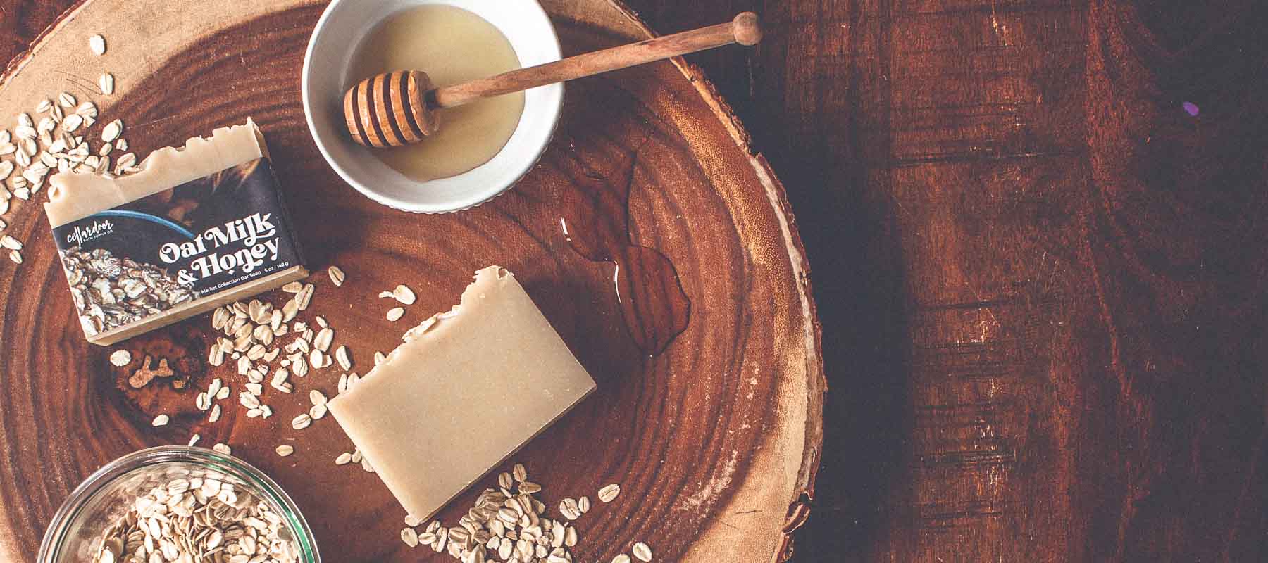 Two bars of a soap names Oat Milk & Honey sit on a wood background surrounded by oats and honey.