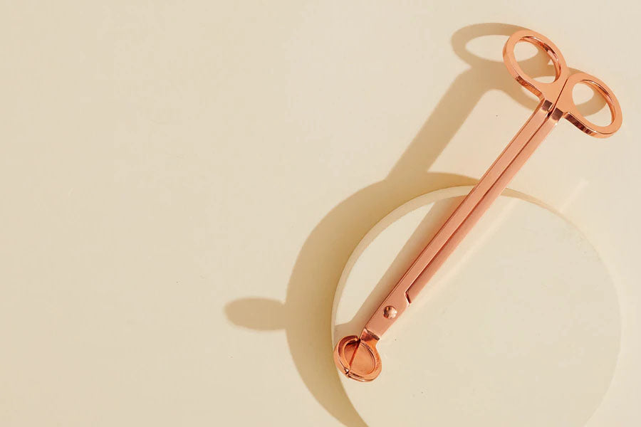 A rose gold colored candle wick trimmer rests on a cream colored background.