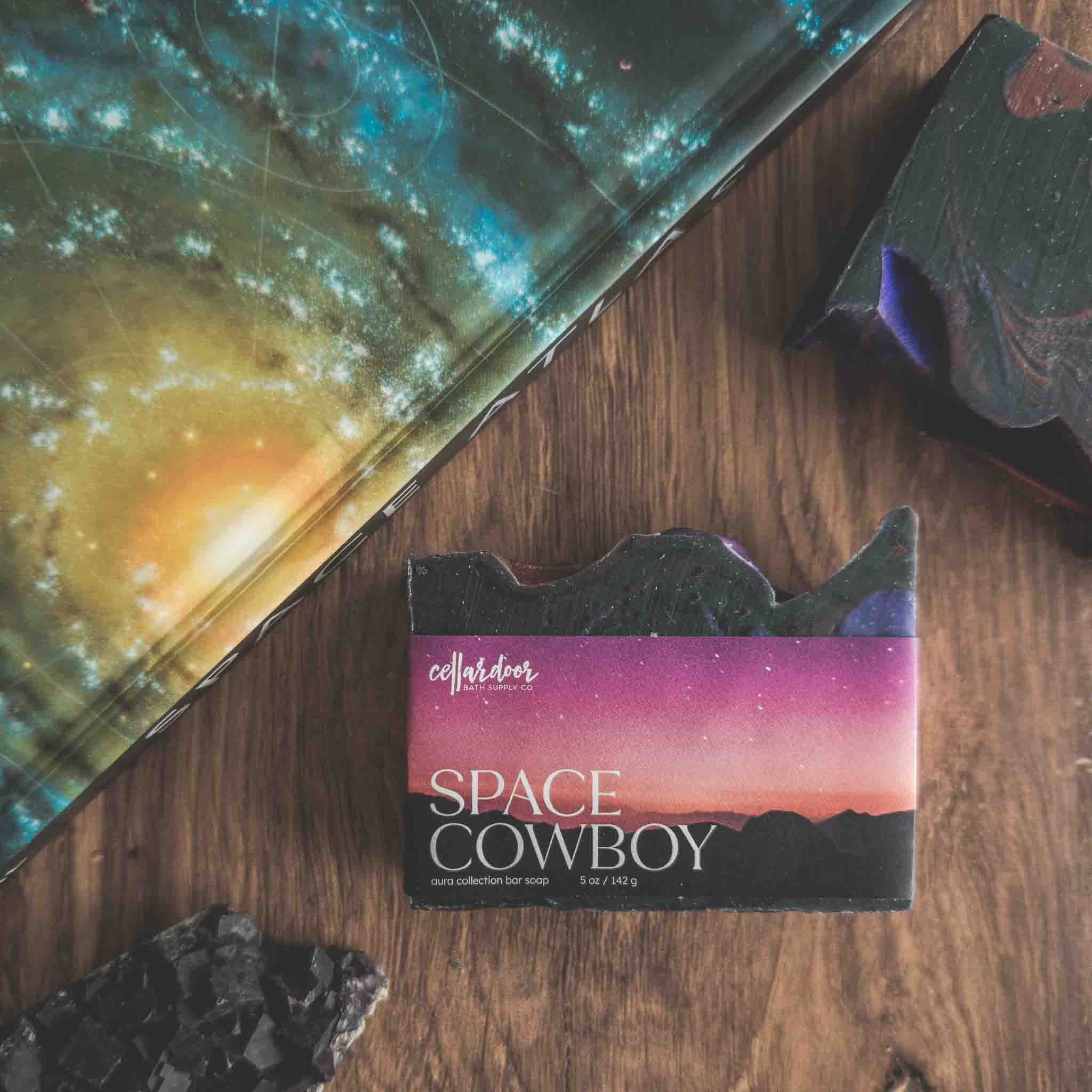 Soap bar with 'Space Cowboy' packaging on a wooden surface next to a galaxy-themed book