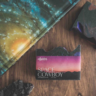 Soap bar with 'Space Cowboy' packaging on a wooden surface next to a galaxy-themed book