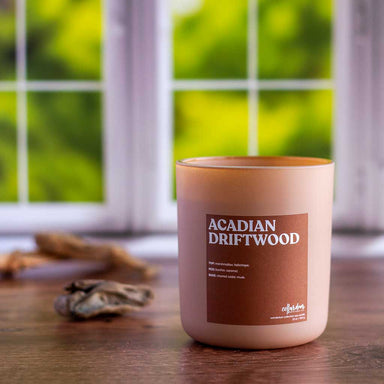 A candle named Acadian Driftwood in a brown jar sits by a windowsill.