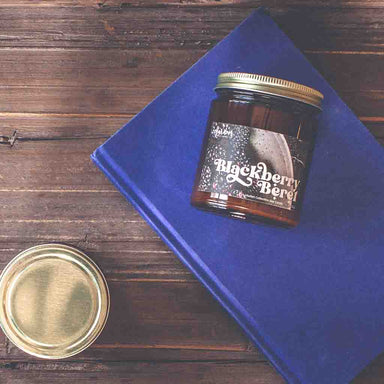 A candle named 'Blackberry Beret' sits on a blue book set against a wooden background.